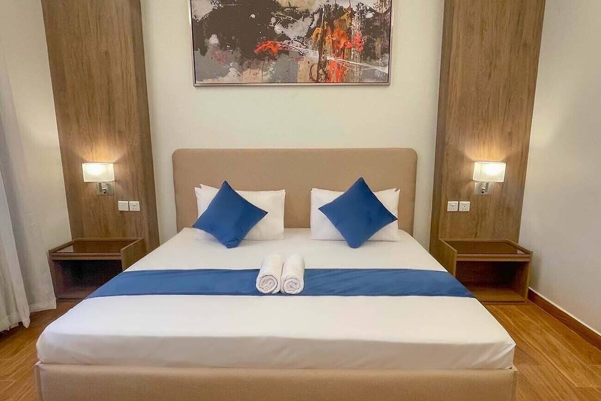 A neatly made bed with a blue accent pillow, two bedside lamps, and a painting above it.