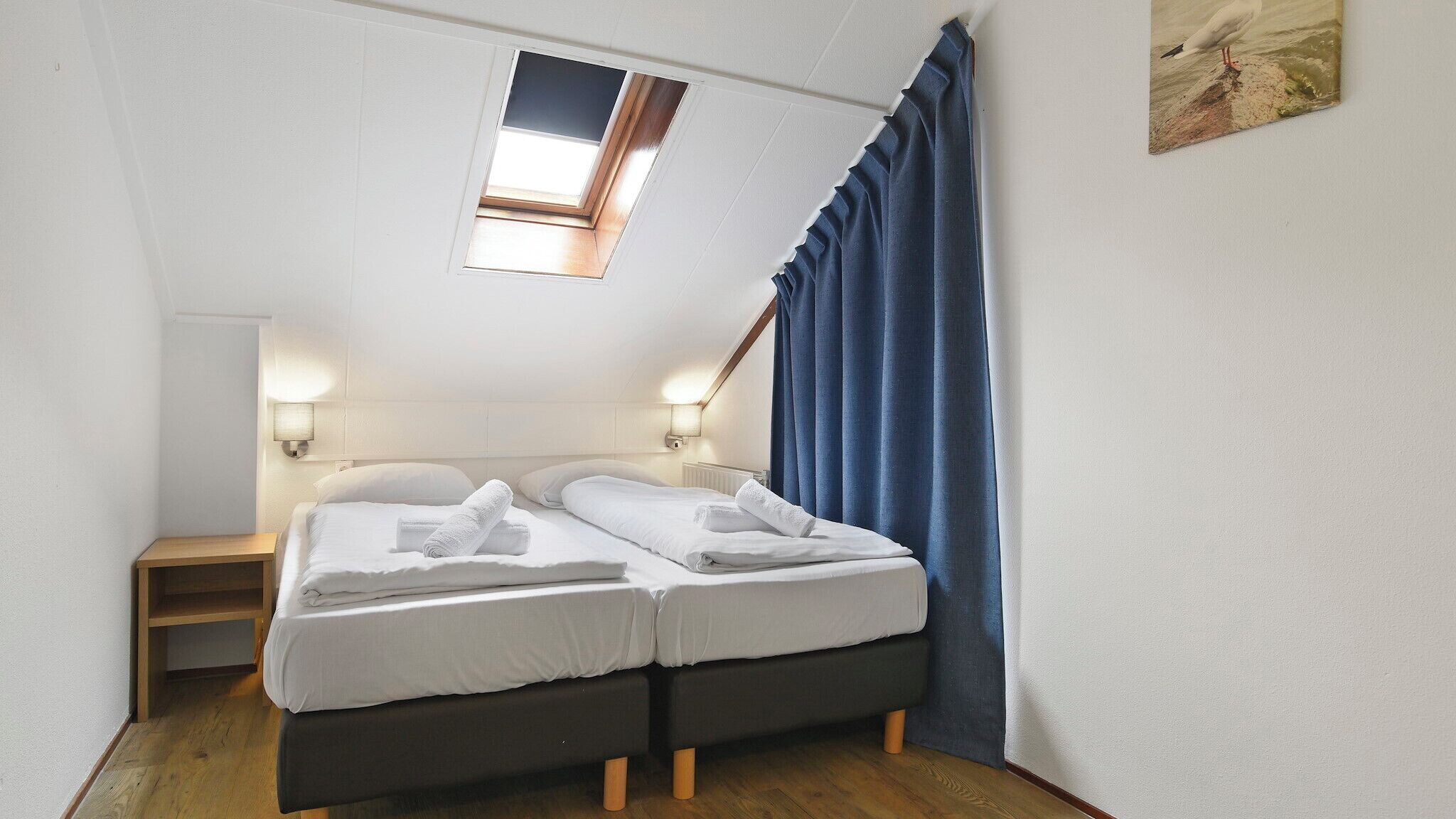 A small, neatly made twin bed with white linens and towels, a small bedside table, a wall-mounted lamp, and a window with a view of the sky.