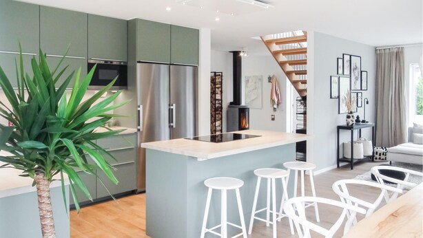 Una cocina moderna con armarios verdes, una isla central con taburetes, una estufa de leña y una escalera que sube a un nivel superior.
