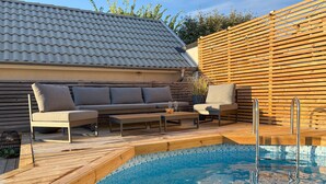 Un área junto a la piscina con una terraza de madera, un conjunto de sofás y una piscina con mosaicos azules.