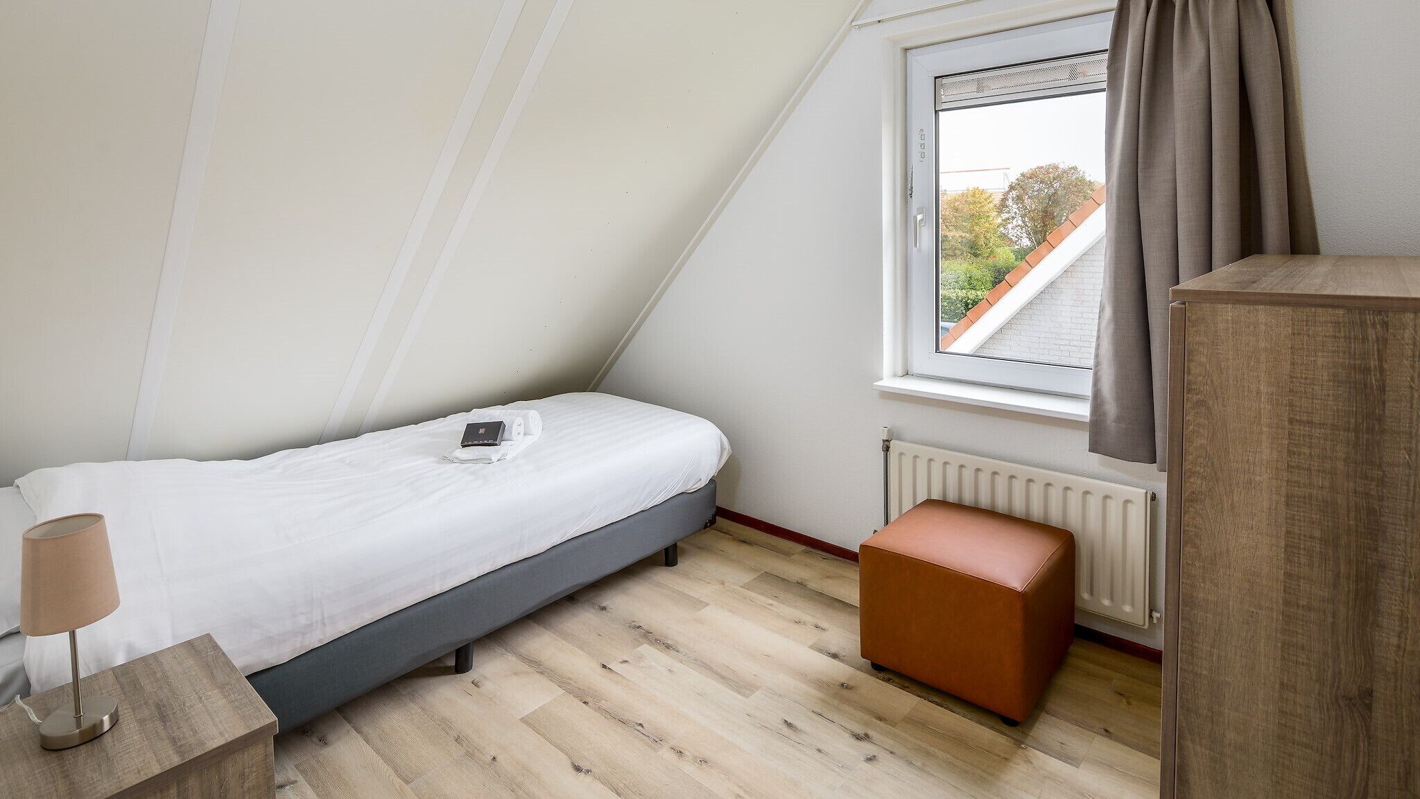 Ein kleines, ordentlich eingerichtetes Schlafzimmer mit einem Einzelbett, einem Nachttisch mit Lampe, einem Fenster mit Vorhängen und einer Heizung.