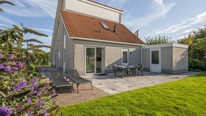 Una casa moderna de dos pisos con techo de tejas, un patio con mobiliario exterior y un césped bien cuidado.
