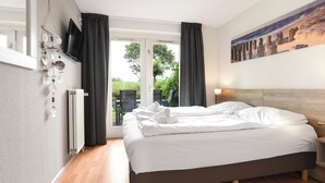 A hotel room with a bed, a television mounted on the wall, a painting, and a view of the outdoors. - Bungalow for 8 persons (Brouwershaven)