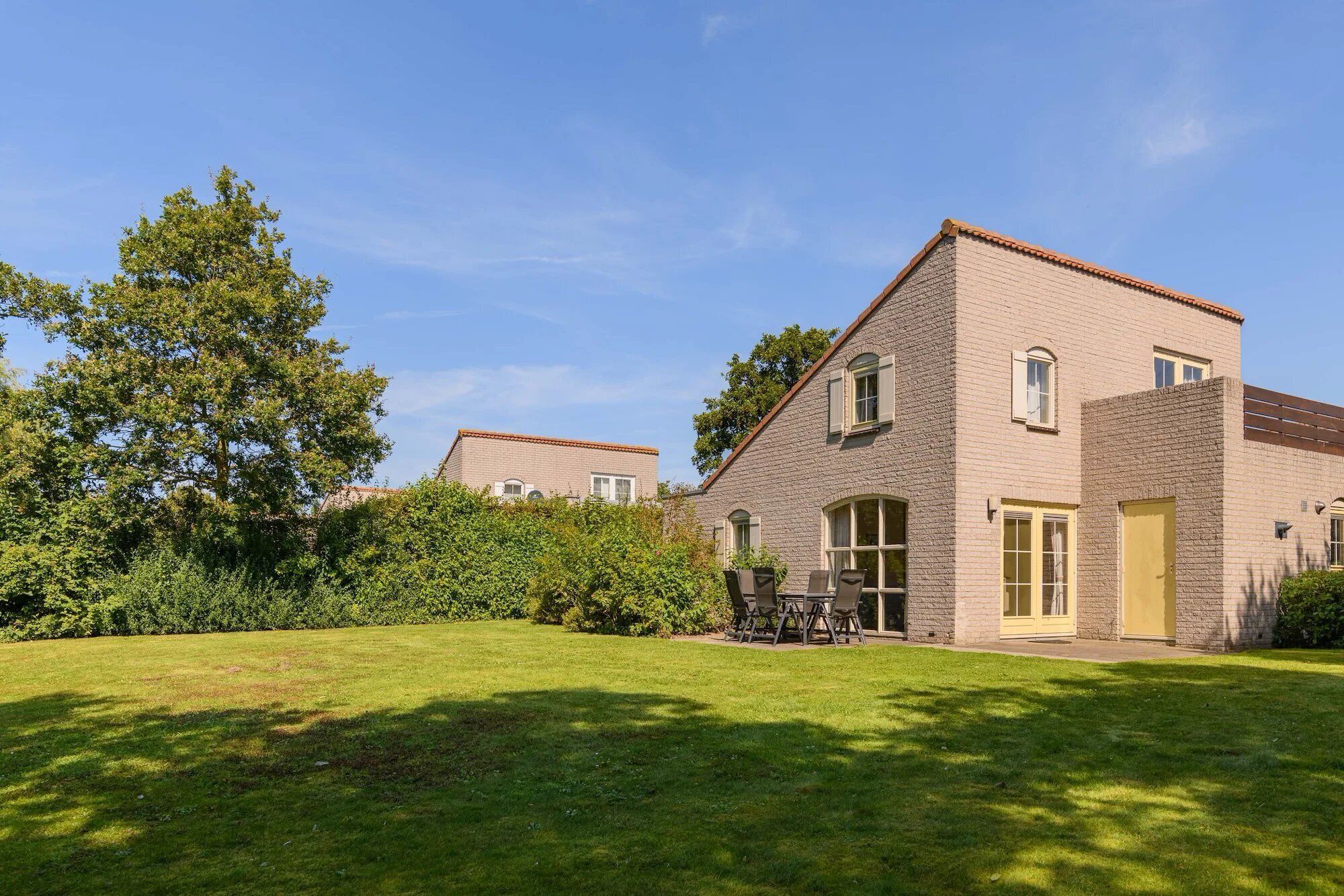 Une maison à deux étages, avec une façade en briques, un toit en tuiles et un jardin devant, comprenant une pelouse et une table de patio.