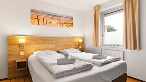 A hotel room with two beds, a wooden headboard, wall-mounted lamps, a painting of a beach, and a window with curtains.