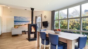 A modern living room with a fireplace, a dining area with blue chairs, a white sofa, and a large window with a view of trees and a beach. - Bungalow for 6 persons with sauna (Brouwershaven)