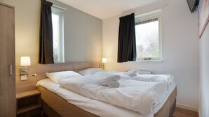A hotel room with a bed, two bedside lamps, a nightstand, a window with curtains, and a mirror. - Bungalow for 6 persons with sauna (Brouwershaven)