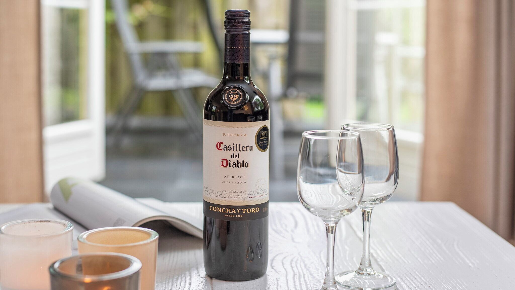 Una botella de vino Merlot Casillero del Diablo, dos copas de vino vacías y tres candelabros sobre una mesa.