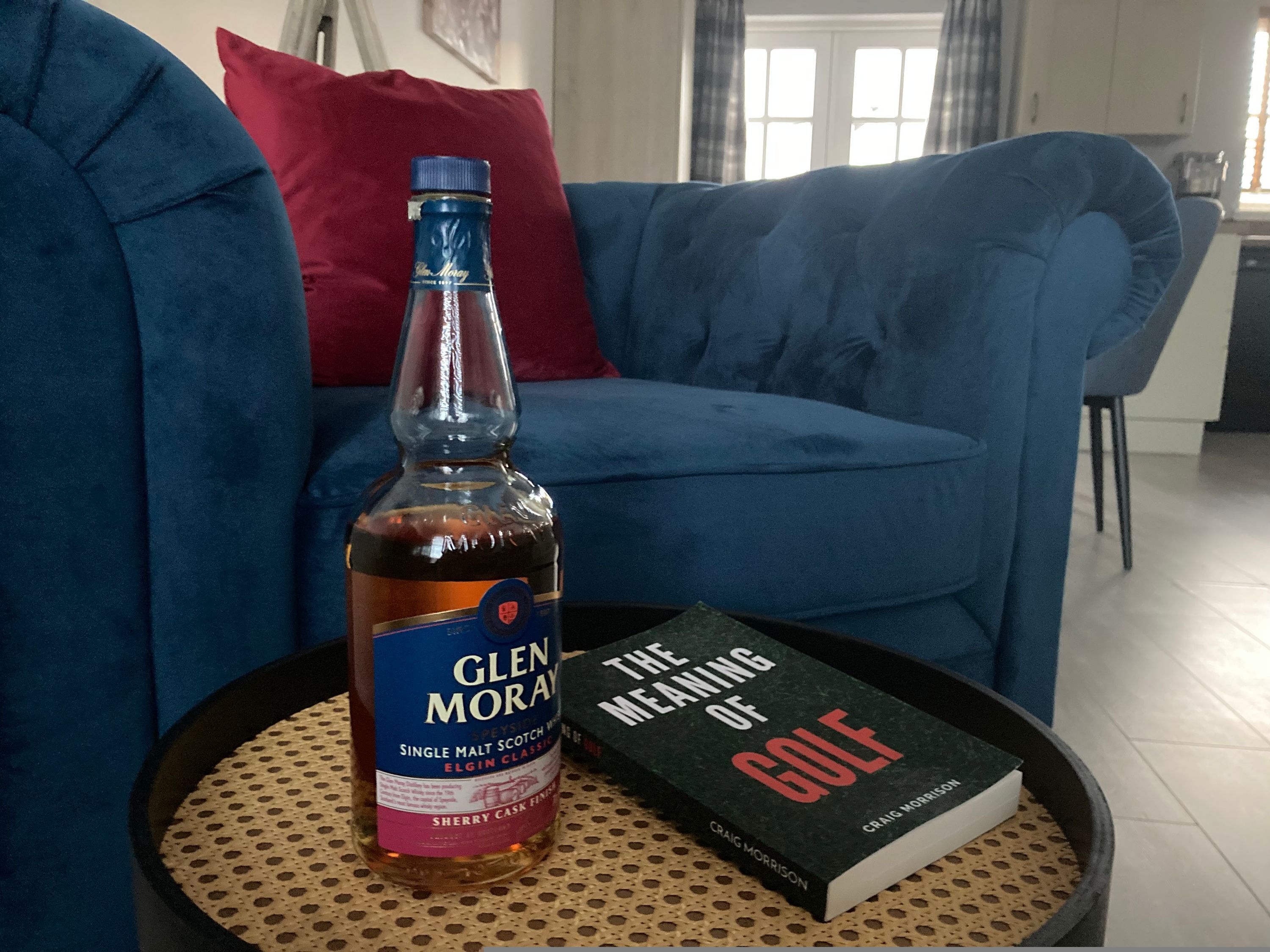A bottle of Glen Moray Single Malt Scotch Whisky and a book titled 'The Meaning of Golf' on a round table.