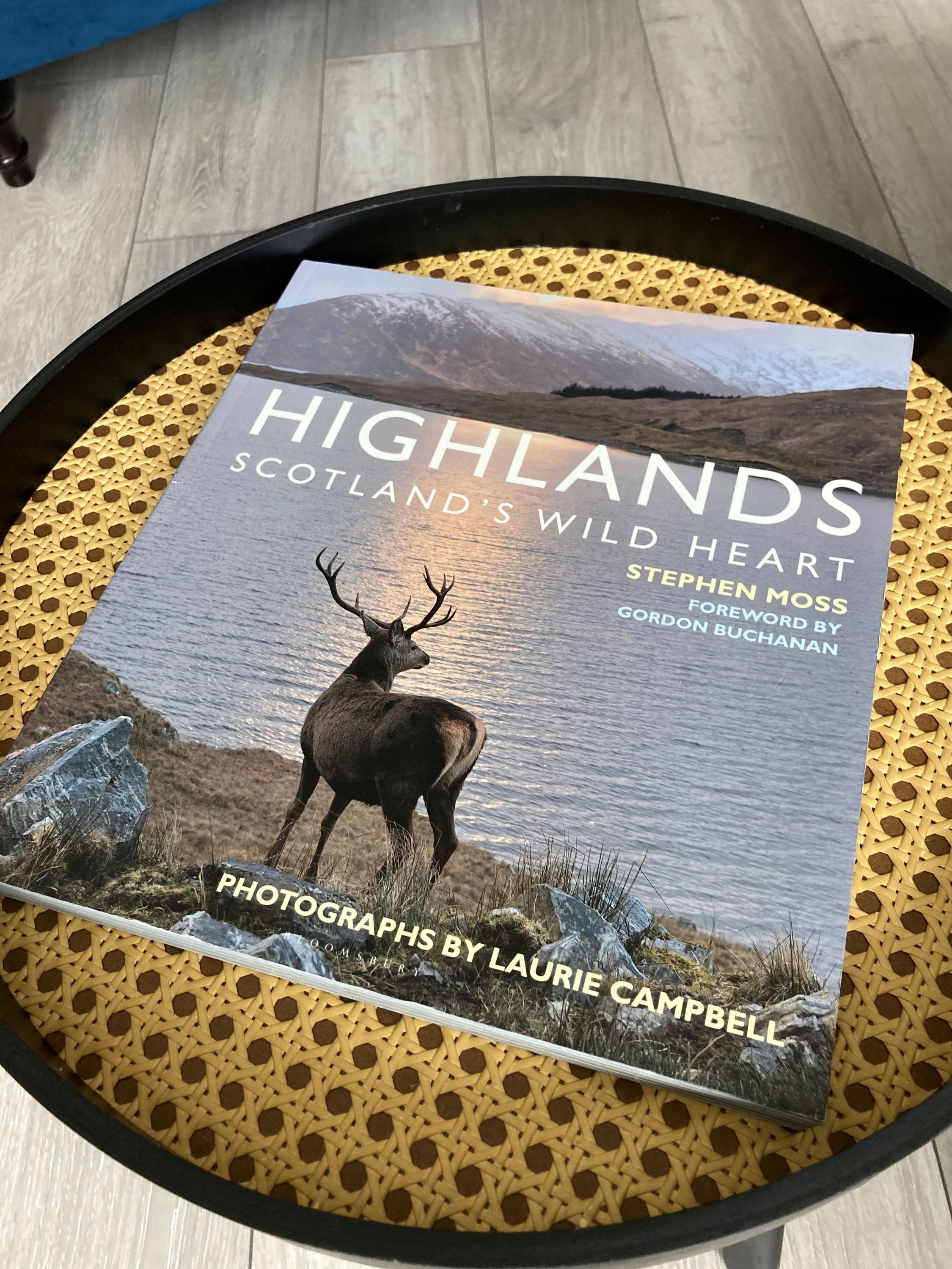 A book titled "Highlands: Scotland's Wild Heart" by Stephen Moss with a foreword by Gordon Buchanan, placed on a round tray with a woven pattern.