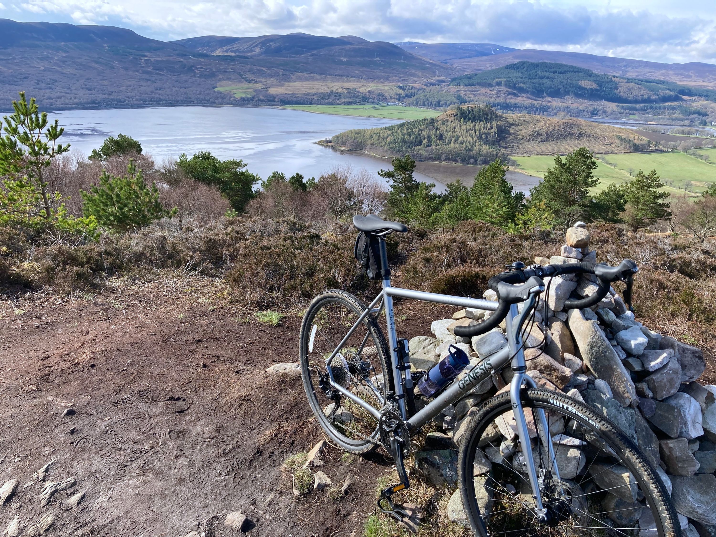 Ein Mountainbike auf einem felsigen Vorsprung mit Panoramablick auf einen See und Hügel im Hintergrund.