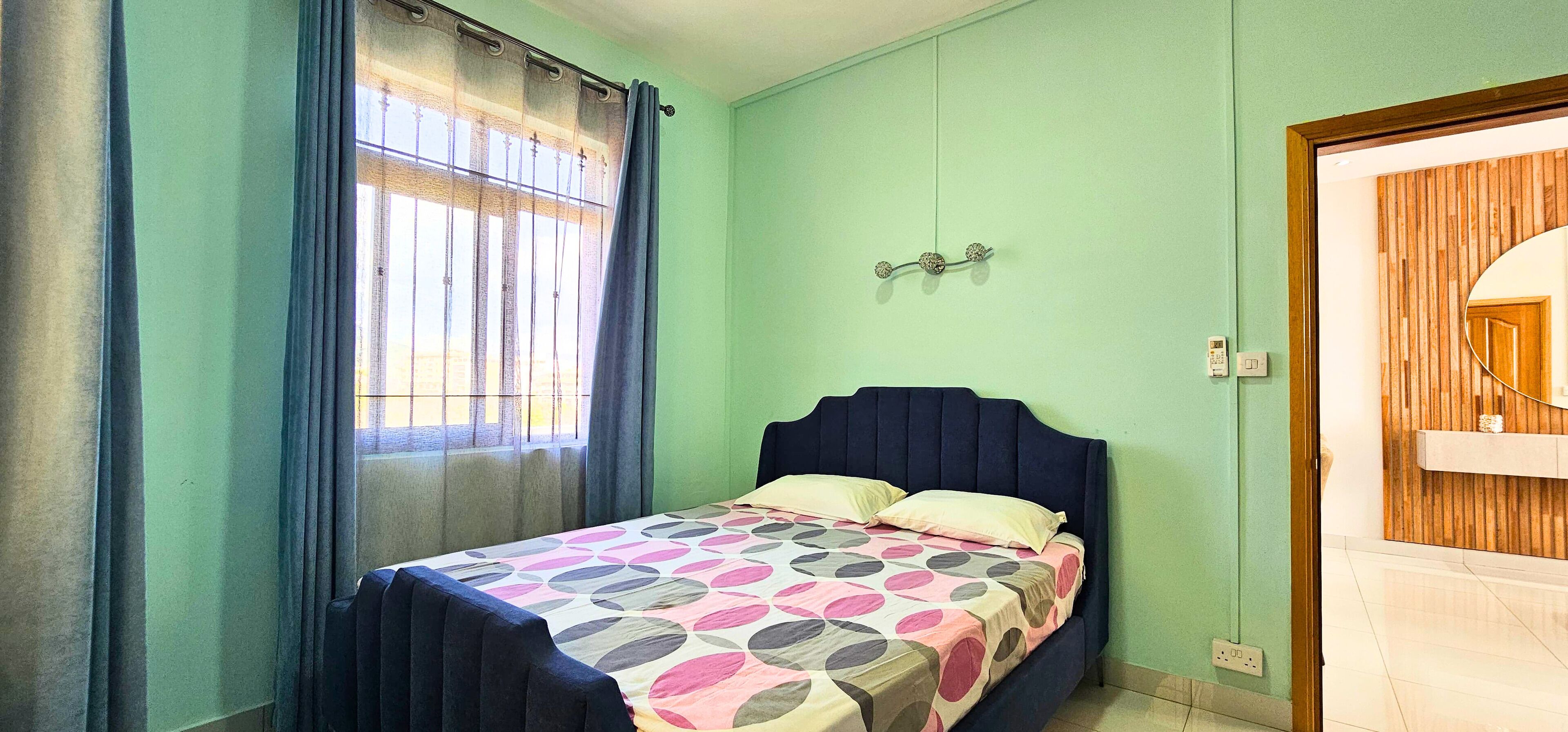 A bedroom with a bed, a window with curtains, a wall-mounted light fixture, and a wooden door.
