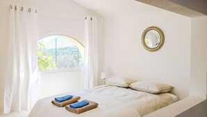 Une chambre à coucher avec un lit, des rideaux blancs, une fenêtre donnant sur un beau paysage et un miroir rond accroché au mur.