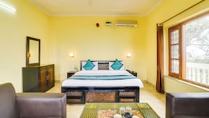 Habitación de hotel con cama, dos sillas, una mesa con objetos, aire acondicionado y ventana con cortinas.