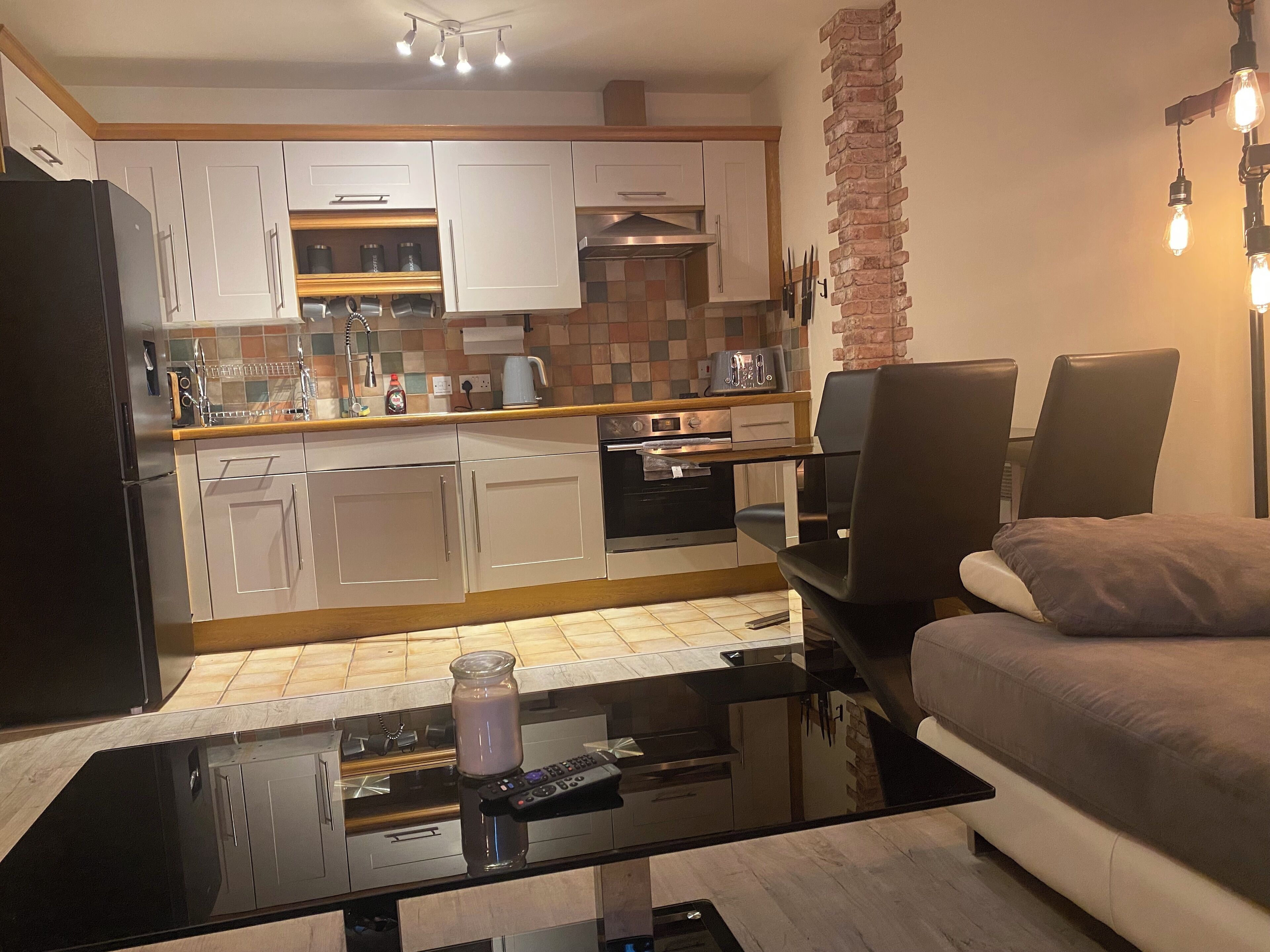 A modern kitchen with white cabinets, a black refrigerator, and a coffee machine. There's a black table with a remote control and a glass jar. A brown sofa and a black chair are in the living area.