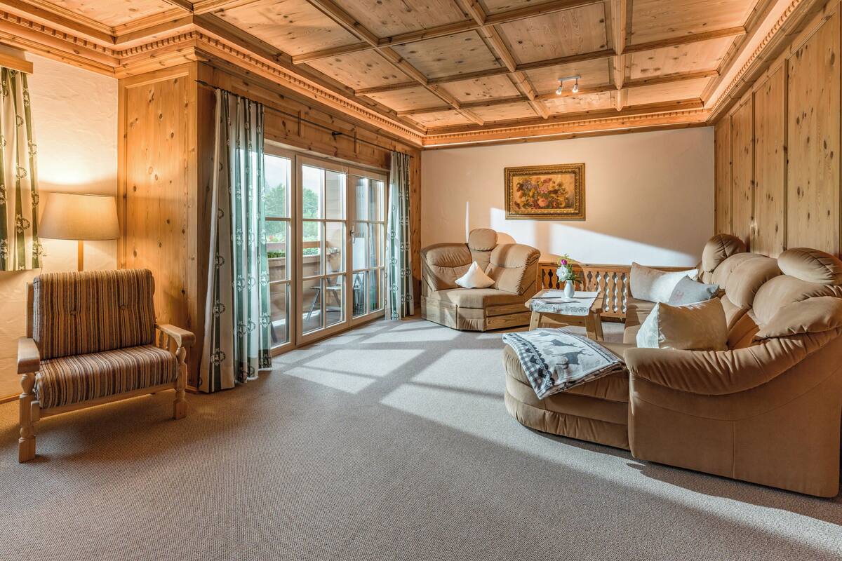 A cozy room with a wooden ceiling, a large window, a sofa, a chair, and a table with a vase.