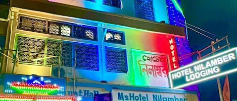 A multi-story building with colorful neon lights, a sign for 'M/s Hotel Nilamber', and motorcycles parked outside.