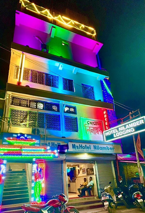 A multi-story building with colorful neon lights, a sign for 'M/s Hotel Nilamber', and motorcycles parked outside. - Hotel Nilamber By GRB (Lanka)