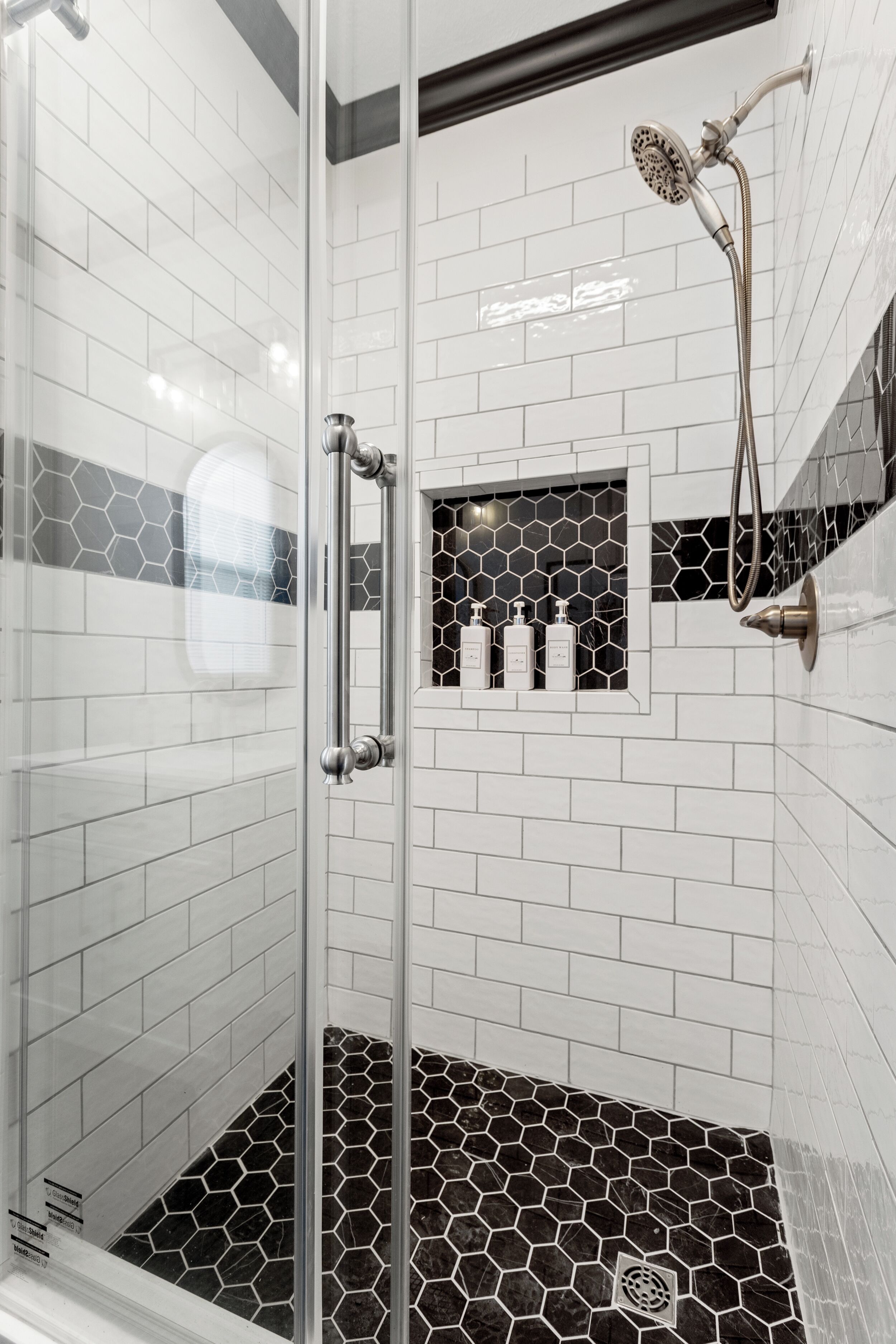 A glass-enclosed shower with a black and white tiled floor and a hexagonal pattern on the lower wall.