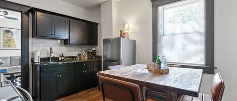 A kitchen with dark cabinets, a wooden dining table, and a window with blinds.
