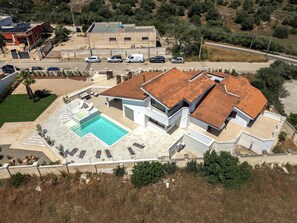 Vista aérea de una casa moderna con piscina, varios autos estacionados afuera y un paisaje montañoso al fondo.