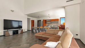 A modern living room with a large flat-screen TV, a sectional sofa, and a dining area with a kitchen in the background.