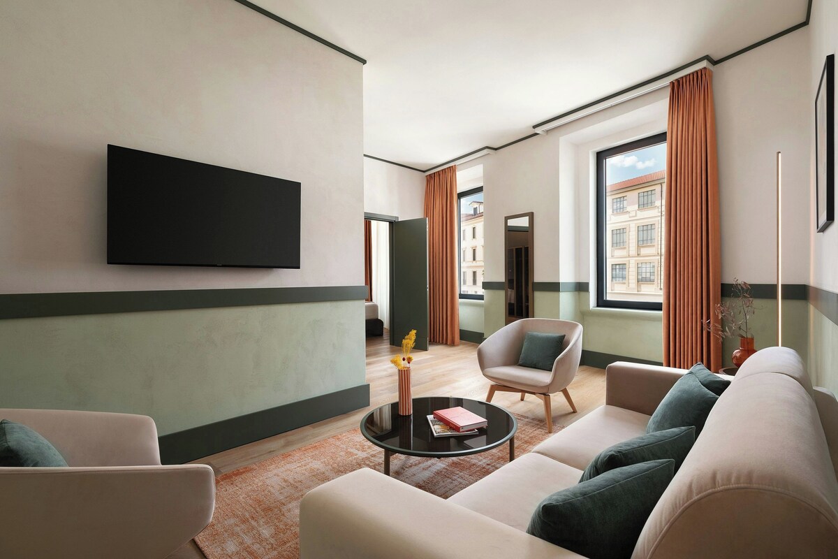 A modern hotel room with a flat-screen TV mounted on the wall, a sofa set, a coffee table, and a view of the city through large windows.