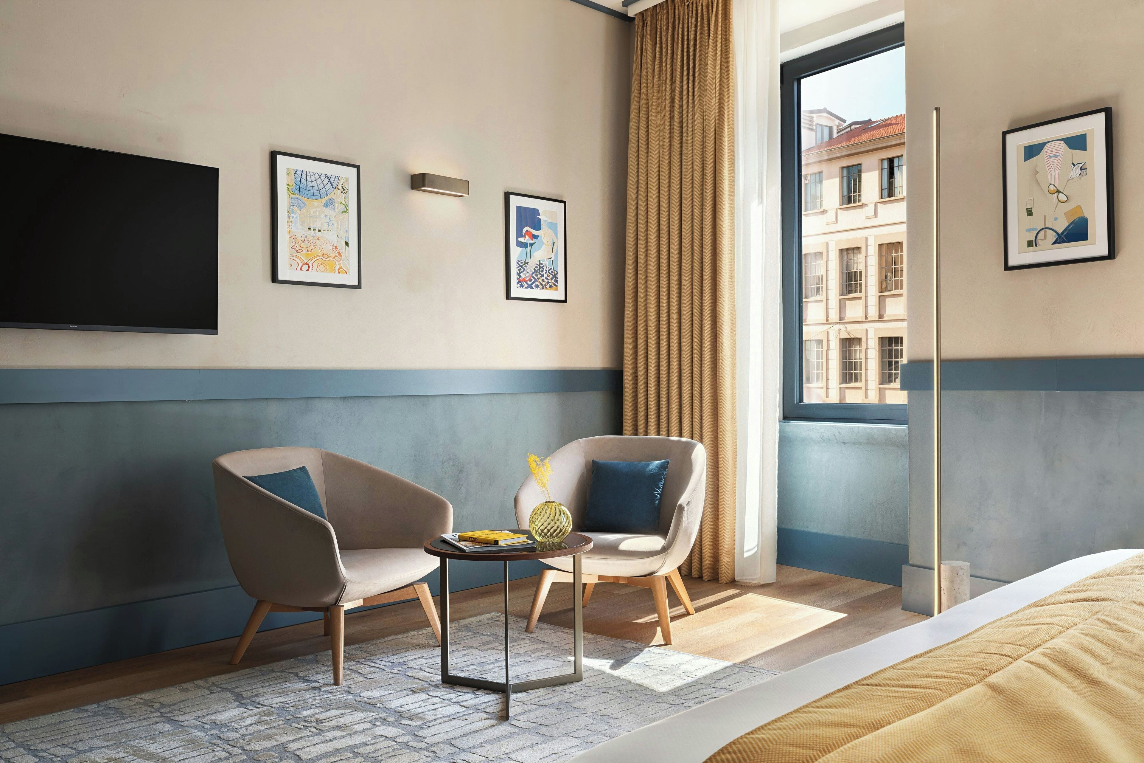 A hotel room with two armchairs, a small table with a vase, a flat-screen TV mounted on the wall, and a window with curtains.