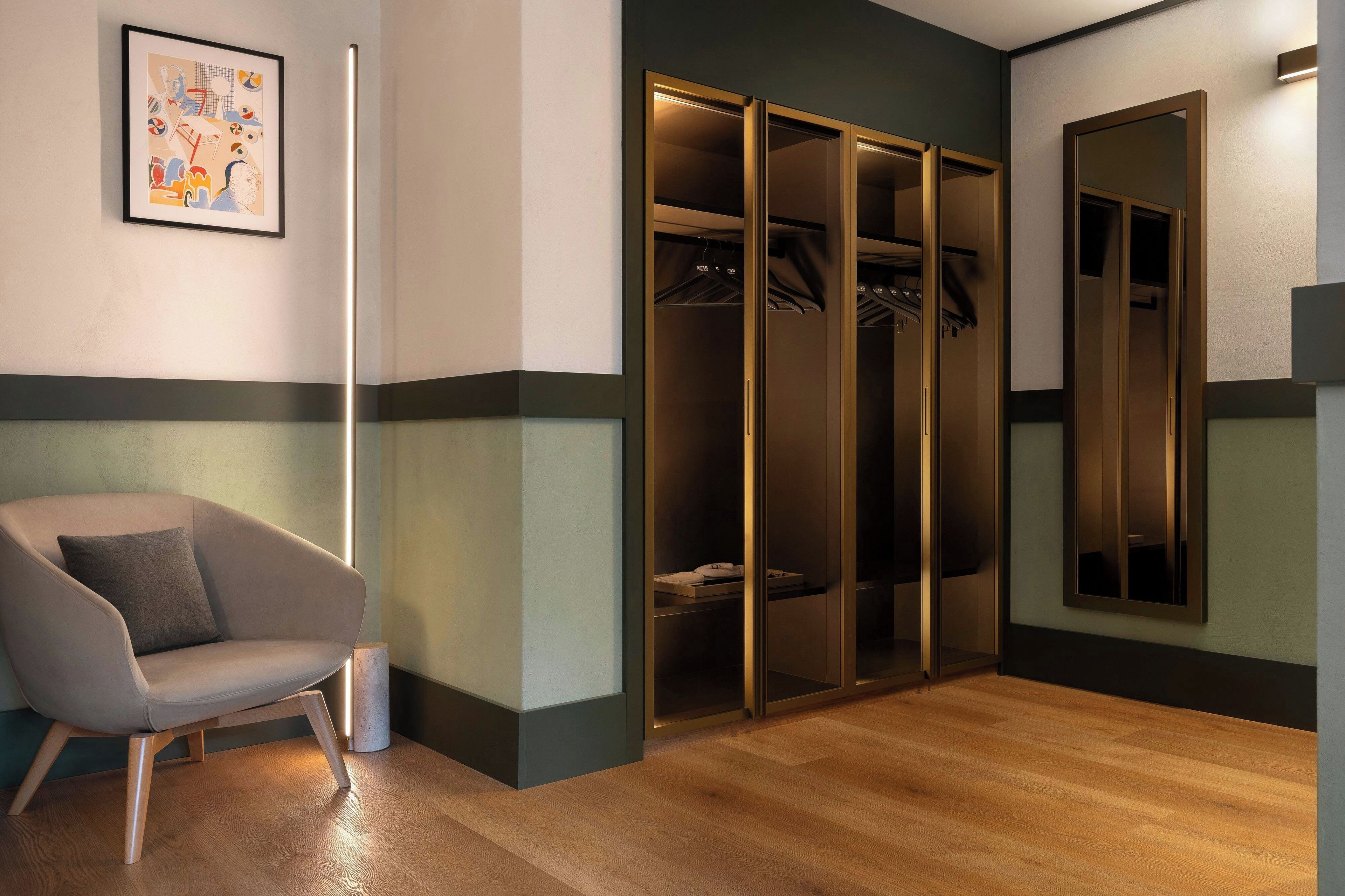 A modern hotel room with a wooden floor, a grey armchair, a large mirror, and a walk-in closet with hanging clothes.