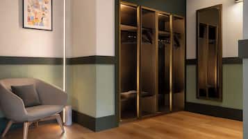 A modern hotel room with a wooden floor, a grey armchair, a large mirror, and a walk-in closet with hanging clothes.