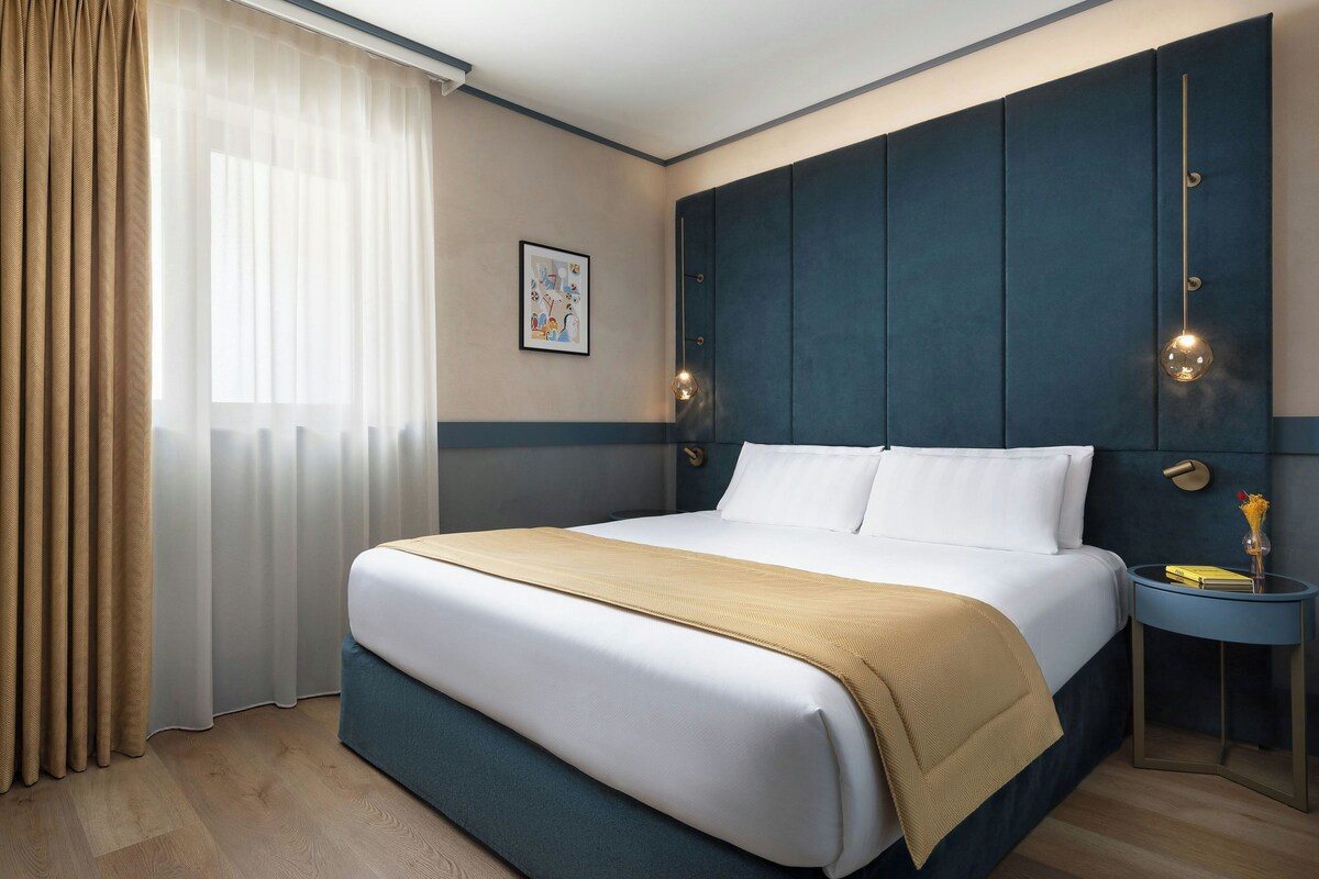 A hotel room with a large bed, a bedside table, a nightstand, and a wall-mounted light fixture.