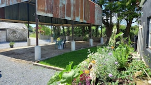 Un espacio de estar al aire libre de estilo rústico, con estructura metálica, edificio de piedra y un jardín con diversas plantas y flores.