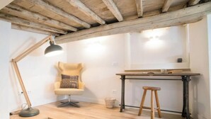 A cozy room with a wooden ceiling, a chair with a cushion, a desk with a stool, and a lamp.