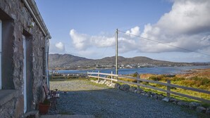 A rustic stone building with a gravel driveway leading to a wooden fence overlooking a coastal landscape with mountains in the distance. - Bridies Holiday Cottage Roundstone (Inishnee)