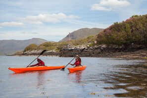 Two people kayaking in orange kayaks on a calm body of water with a rocky shoreline and mountains in the background. - Bridies Holiday Cottage Roundstone (Inishnee)