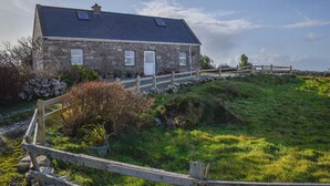 A stone house with a sloped roof, a wooden fence, and a grassy hill in the foreground. - Bridies Holiday Cottage Roundstone (Inishnee)