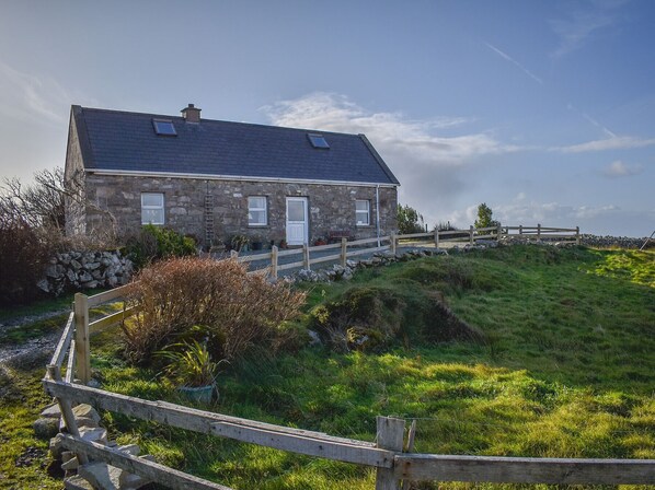 A stone house with a sloped roof, a wooden fence, and a grassy hill in the foreground. - Bridies Holiday Cottage Roundstone (Inishnee)