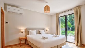 Een slaapkamer met een groot bed, bedlampen, een airconditioner, een klein tafeltje en uitzicht op groen buiten het raam.