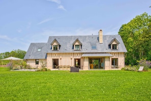 Une grande maison avec un toit en pente, de nombreuses fenêtres et un vaste jardin à l’avant.
