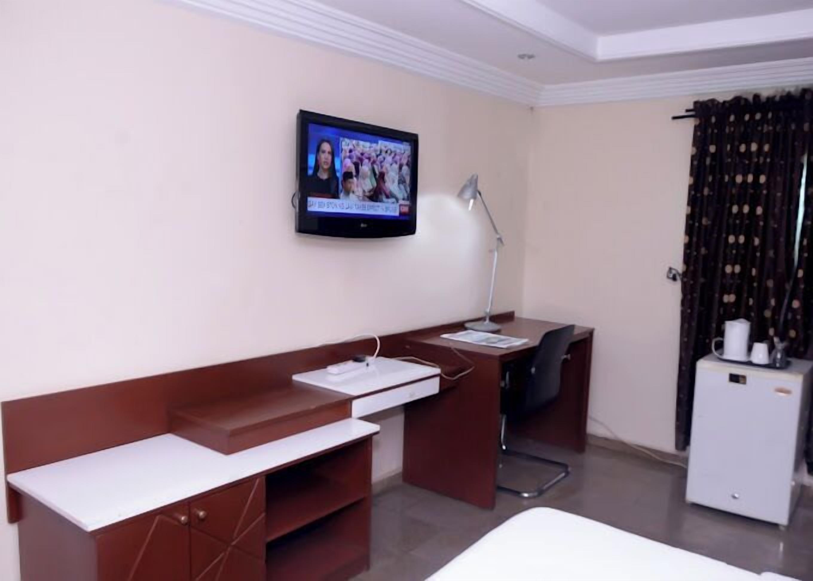 A hotel room with a desk, a television mounted on the wall, a desk lamp, and a small refrigerator.