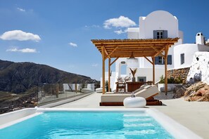 A modern pool area with a wooden pergola, white walls, and a clear view of the surrounding landscape. - Bluewhite Suites & Villa Santorini (Santorini)