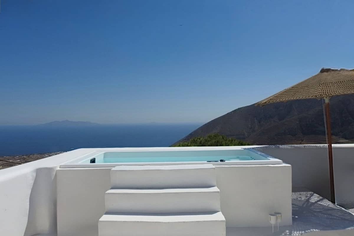 A white infinity pool with steps leading down to a beach, overlooking a mountainous landscape and the sea.