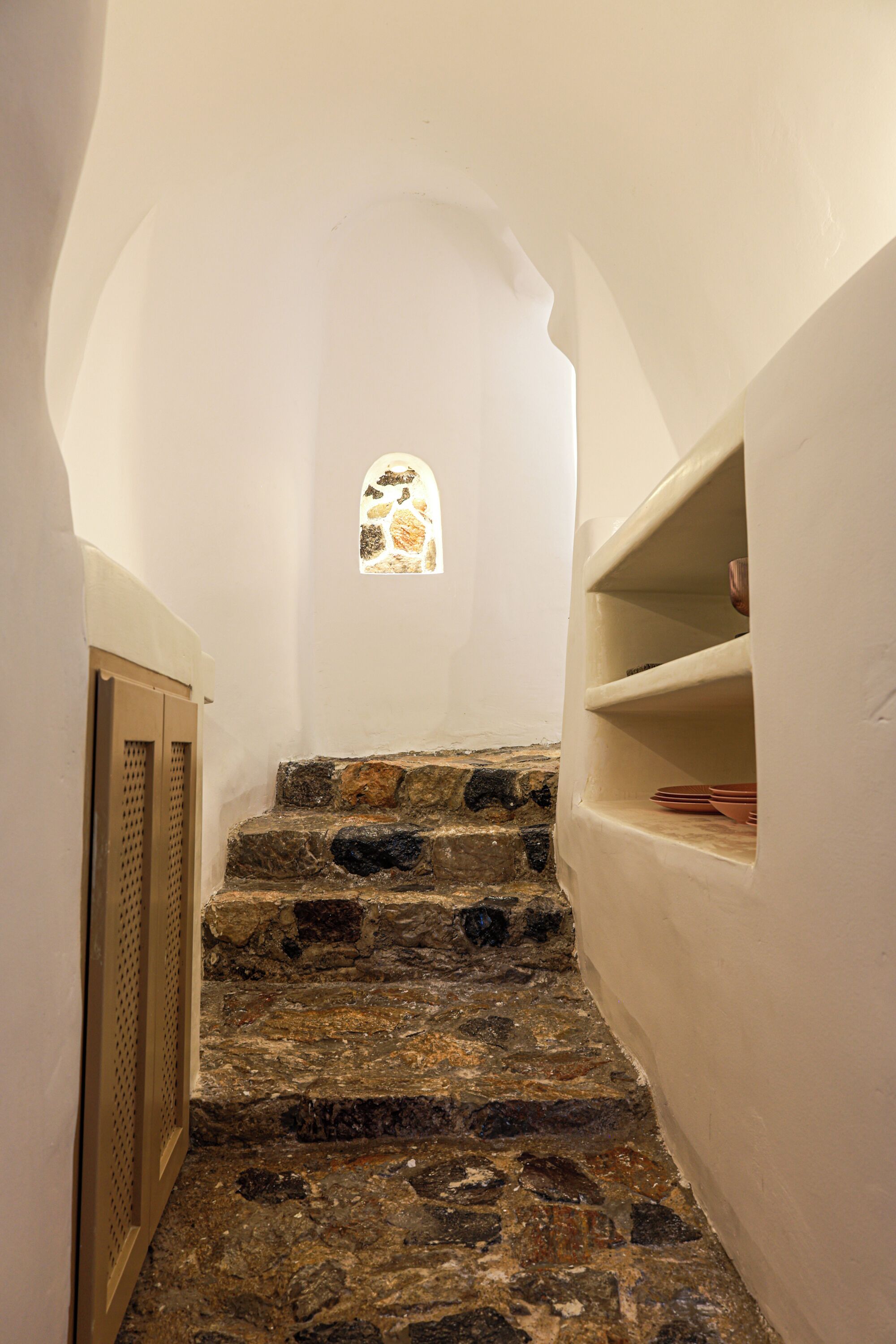 A narrow stone staircase leading to a room with a small stained glass window and shelves with dishes.