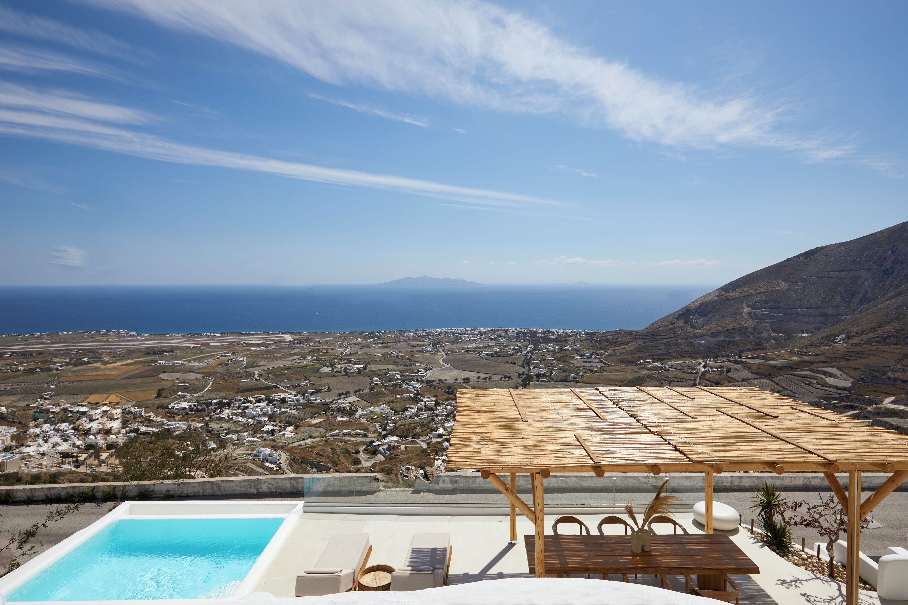 A rooftop terrace with a pool, wooden pergola, and a view of a coastal town and mountains.
