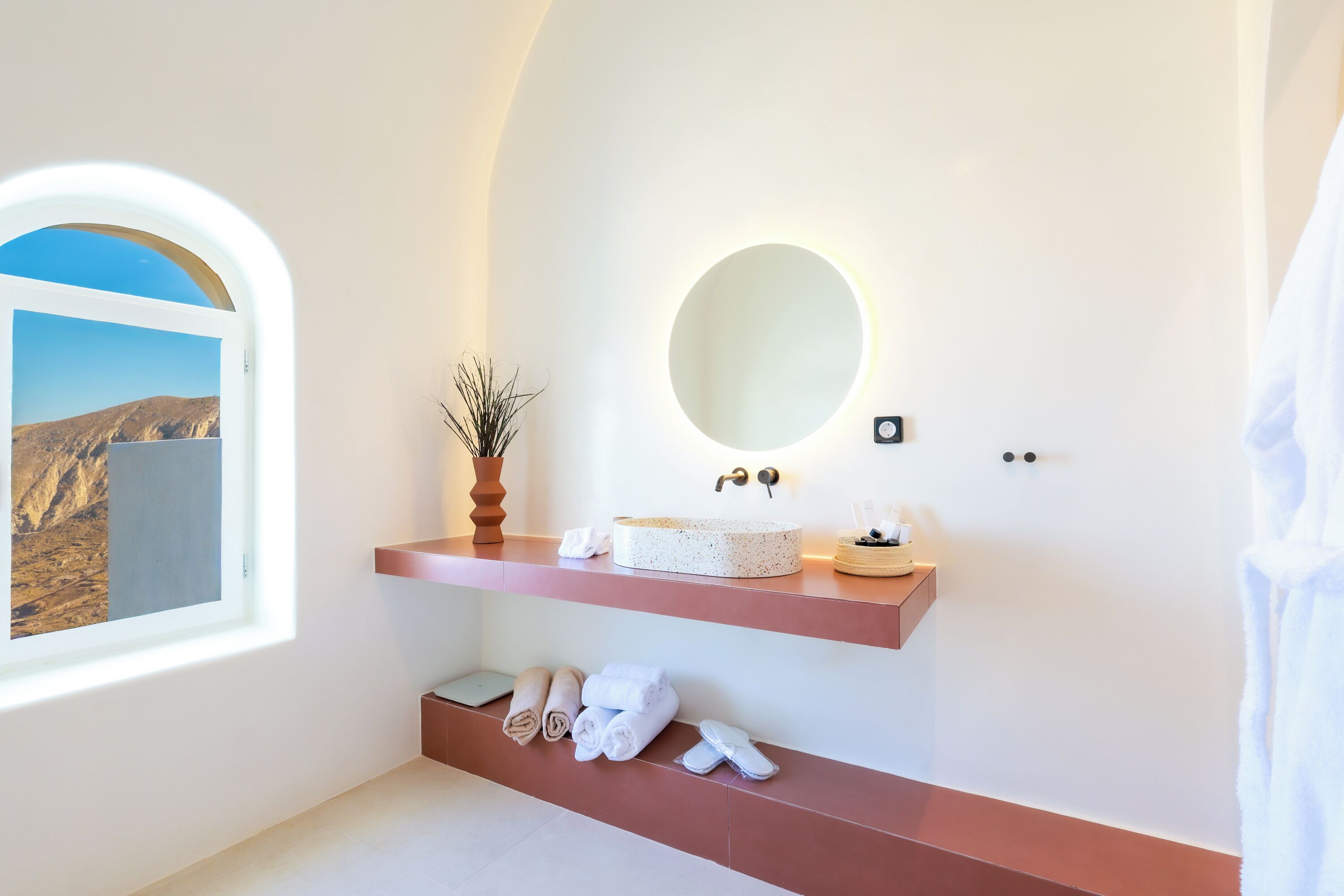 A modern bathroom with a large arched window, a round mirror, and a built-in sink with a towel rack below.