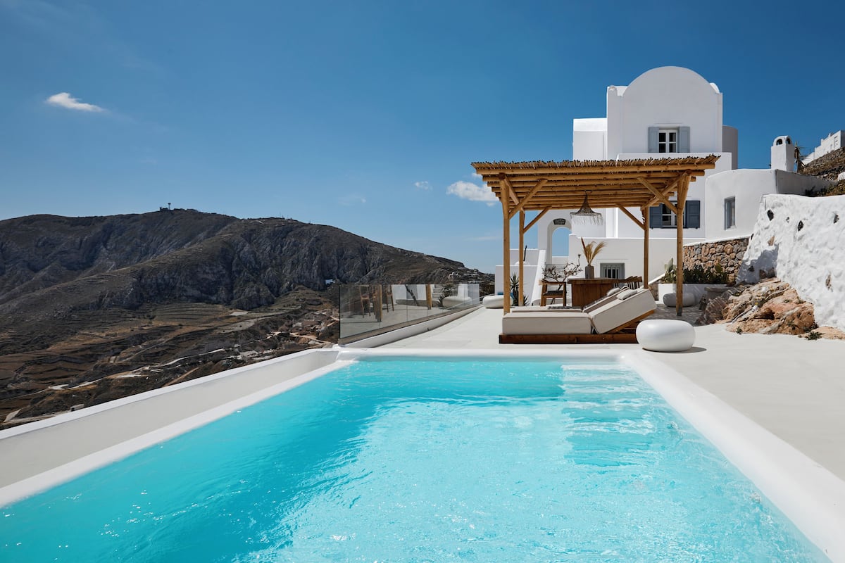 A modern pool area with a wooden pergola, white architecture, and a mountainous backdrop.