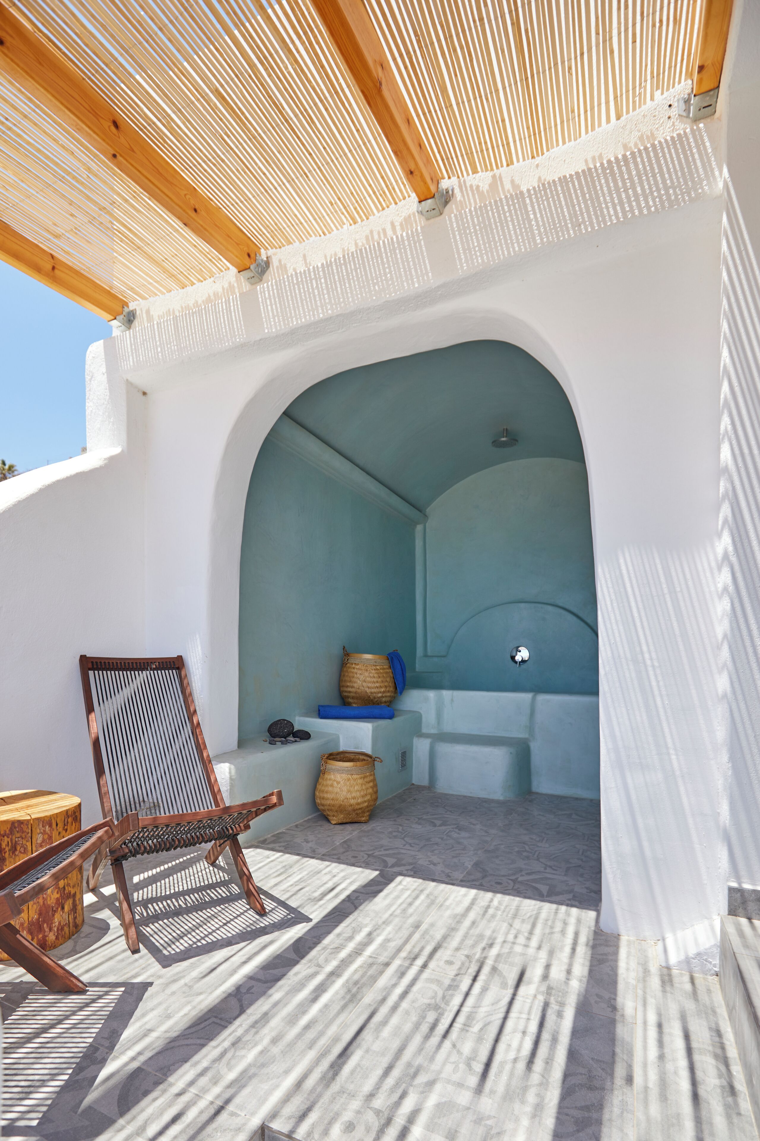 A sunlit outdoor area with a wooden pergola, a stone bench, a wooden chair, and a stone bathtub.