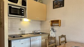 Cocina compacta con microondas, horno, fregadero y lavavajillas. Hay una mesa de comedor de madera con sillas y una pequeña repisa de madera sobre el fregadero.
