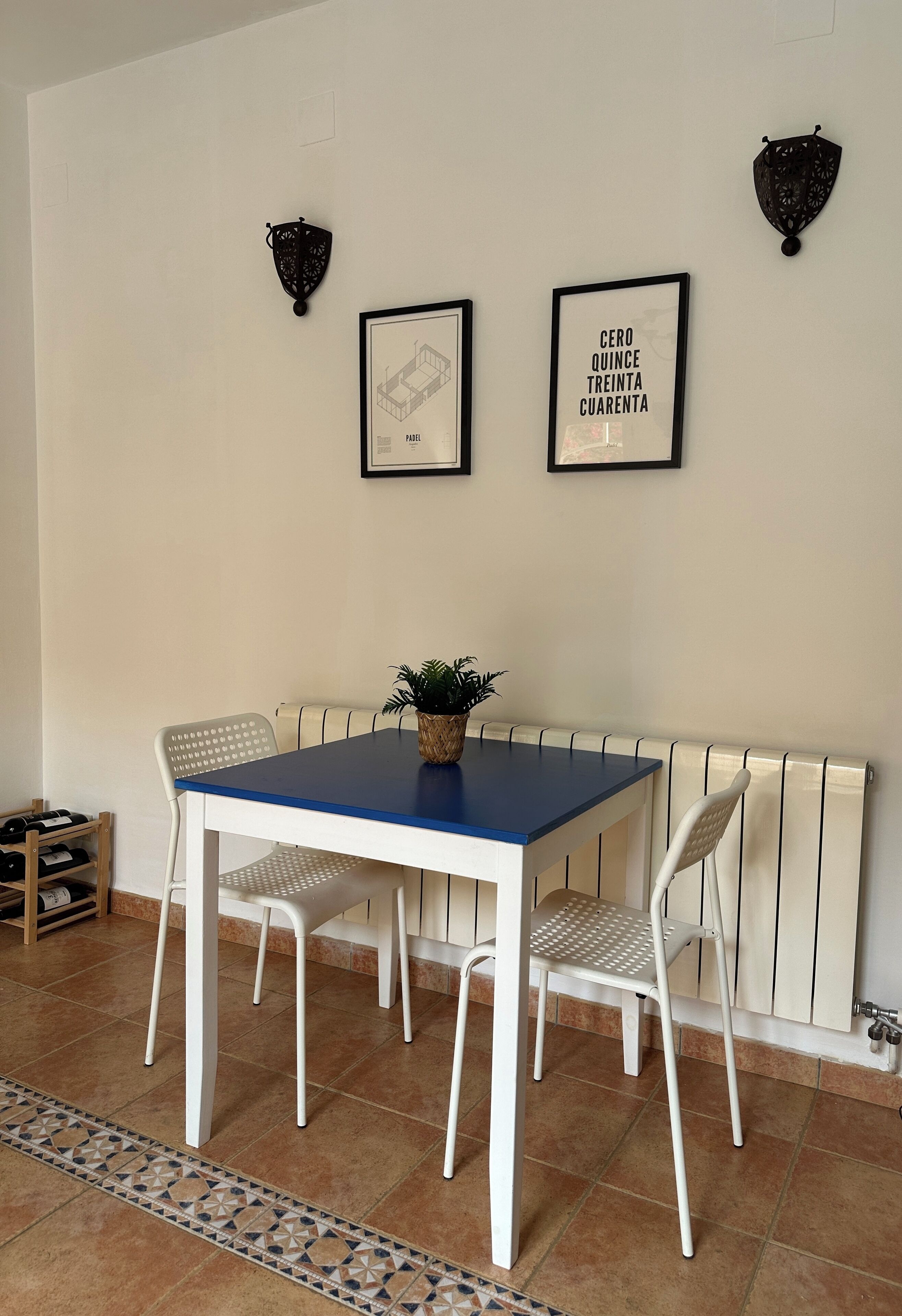 A dining area with a blue table, white chairs, a potted plant, and two framed pictures on the wall.
