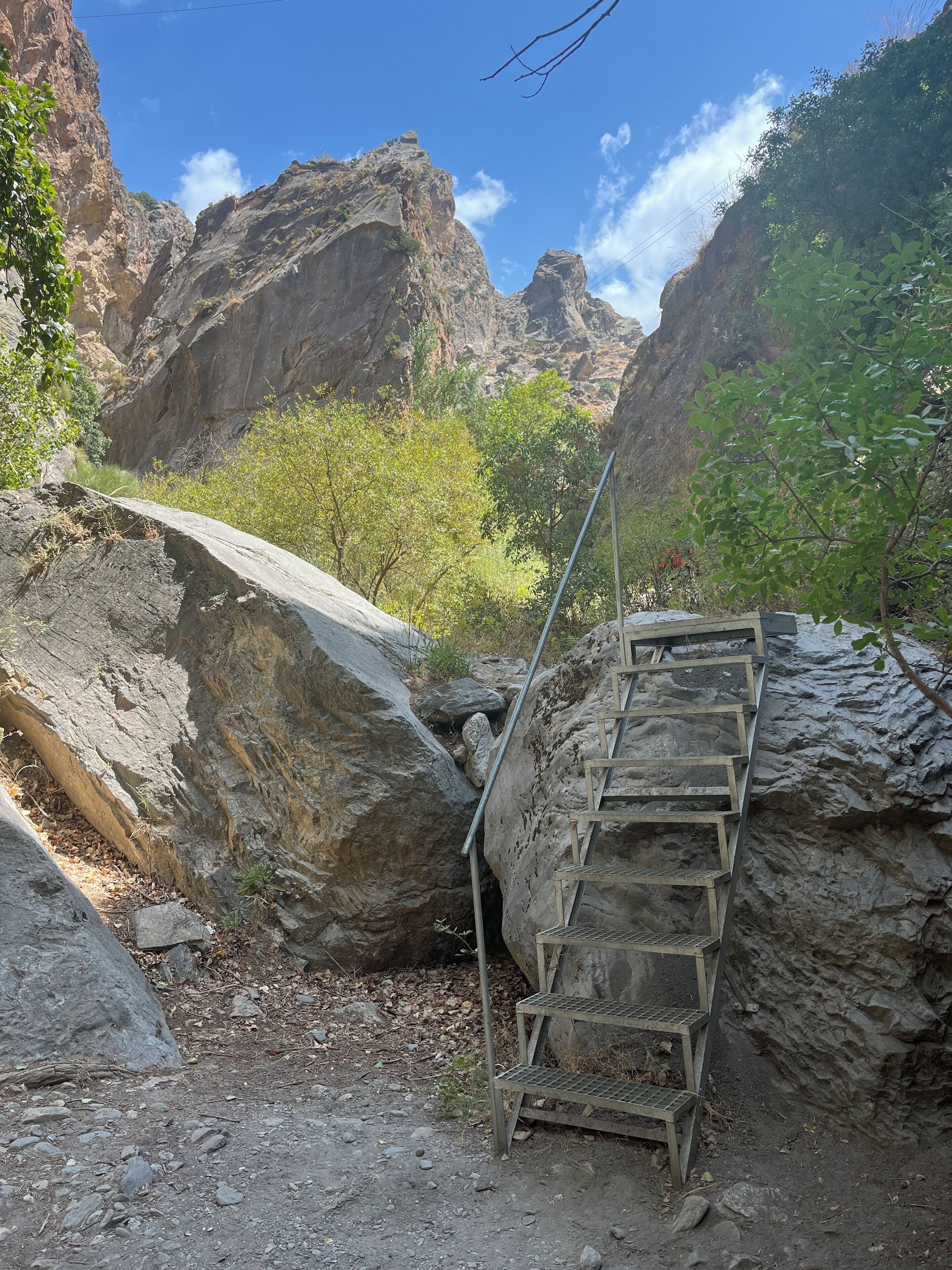 Terreno rocoso con una escalera metálica que asciende por una pendiente pronunciada, rodeado de grandes rocas y vegetación escasa.